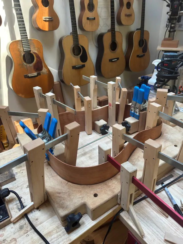 A body mold full of cauls and clamps, vintage guitars hanging behind the bench